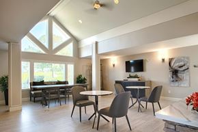 Mountlake Terrace apartment for rent - A modern resident gathering space here at Andorra featuring a high vaulted ceiling with triangular windows, round café tables with upholstered chairs, a long dining table with bench seating near large windows, light wood-style flooring, neutral walls with abstract artwork, wall-mounted television, and soft lighting that creates a bright, welcoming atmosphere.