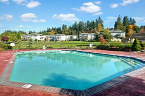 Mountlake Terrace apartment for rent - A refreshing outdoor swimming pool here at Andorra featuring clear blue water surrounded by a spacious red-toned pool deck, metal handrails, and safety fencing, with expansive green lawns, mature trees, and surrounding apartment buildings in the background, creating a serene community setting ideal for relaxing, swimming, and enjoying sunny days.