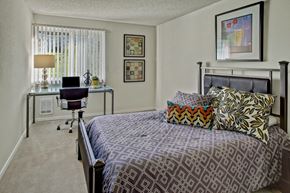 Mountlake Terrace apartment for rent - A comfortable bedroom here at Andorra featuring a neatly made bed with patterned bedding and accent pillows, a metal bed frame, and framed artwork on light-colored walls, complemented by soft carpeting, a window with vertical blinds, and a functional desk setup that creates a quiet space for rest and productivity.