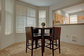 Kirkland apartment for rent - A cozy dining nook here at Aspen Creek featuring a tall square dining table with four upholstered chairs set on soft carpet, surrounded by multiple windows with blinds that bring in natural light, and positioned beside a breakfast bar opening into the kitchen with warm wood cabinetry and clean, neutral finishes.