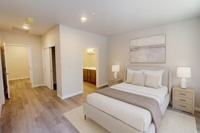 Leavenworth apartment for rent - A serene primary bedroom here at Autumn Haus featuring wide plank wood-style flooring, a neutral upholstered bed with layered bedding, matching nightstands with lamps, and recessed ceiling lighting, with a private ensuite bathroom visible through the doorway, soft neutral walls, and a clean, spacious layout designed for comfort and relaxation.