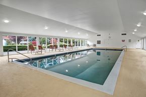 Bellevue apartment for rent - A spacious indoor swimming pool here at Colonial Square featuring a long rectangular pool with stainless steel handrails, a wide pool deck with lounge chairs arranged along floor-to-ceiling windows, smooth light-toned flooring, recessed ceiling lighting, and views of landscaped greenery outside that create a calm and inviting atmosphere.