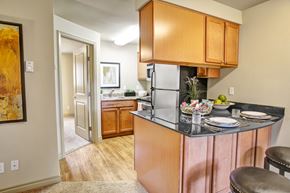 Bellevue apartment for rent - A compact kitchen here at Colonial Square featuring warm wood cabinetry, dark countertops with bar seating, and a small breakfast bar set with plates and glassware, complemented by wood-style flooring, a stainless-look refrigerator, a nearby sink area with additional cabinets, and soft overhead lighting that creates a functional and welcoming cooking space.