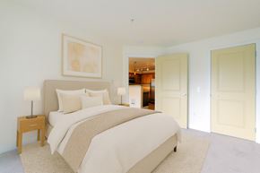 Shoreline apartment for rent - A calm bedroom here at Forest Hills featuring soft neutral walls, light carpet, and a large bed with layered neutral bedding. Matching nightstands with lamps sit on either side, while framed wall art adds a subtle decorative touch. An open doorway leads toward the kitchen, creating a bright and inviting layout.