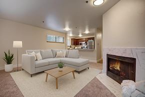 Tukwila apartment for rent - A modern living room here at Forest Greens featuring beige carpet, soft neutral walls, and an open layout that flows toward the kitchen. The space is staged with a light sectional sofa, neutral pillows, a wooden coffee table, a floor lamp, and greenery, while a tiled fireplace adds a cozy focal point.