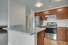 Tukwila apartment for rent - A spacious open kitchen here at Forest Greens featuring rich wood cabinets, light stone-style countertops, and a full appliance setup with a refrigerator, range, and hood. A wide breakfast bar overlooks the adjacent living area, while wood-look flooring and ceiling lights create a warm and functional cooking and gathering space.