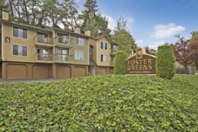 Tukwila apartment for rent - A welcoming exterior view here at Forest Greens showcasing the community monument sign set among manicured greenery, with multi-story apartment buildings in warm neutral tones behind it. The scene features private balconies, garage access below the residences, mature trees, and a landscaped setting that creates a peaceful residential atmosphere.