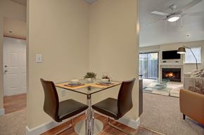 Tukwila apartment for rent - A cozy dining nook here at Forest Greens featuring wood-style flooring and a compact table set for two with modern chairs. The space sits between the entry and living area, offering a clear view toward a carpeted living room with a fireplace, ceiling fan, and sliding glass door that brings in natural light.