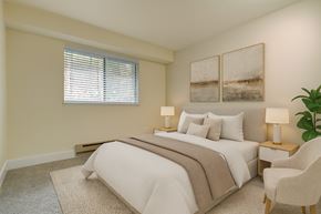 Tukwila apartment for rent - A serene bedroom here at Forest Greens featuring beige carpet and soft neutral walls, styled with a queen bed dressed in white bedding, layered neutral pillows, and a textured throw. Matching bedside tables with lamps flank the bed, abstract wall art hangs above the headboard, and a window with blinds brings in natural light.