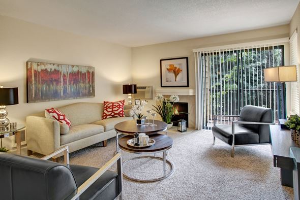 Renton apartment for rent - A stylish living room here at Heritage Hills featuring plush carpet flooring, soft neutral wall tones, and a comfortable seating arrangement with a sofa and accent chairs. The space includes layered round coffee tables, decorative wall art, a tiled fireplace, and a sliding glass door with vertical blinds that opens to a private patio area surrounded by greenery.