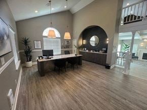 Kent apartment for rent - A welcoming leasing office here at Island Park featuring wood-style flooring, a central desk with computer stations and chairs, and modern pendant lighting overhead. The space includes a curved accent wall with a built-in counter, decorative mirror, greenery, and an open layout that connects to the clubhouse seating area beyond.