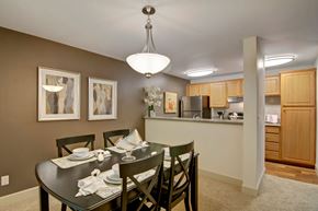 Kent apartment for rent - A welcoming dining area here at Island Park featuring a dark wood dining table set for four beneath a modern pendant light, with framed abstract artwork on an accent wall. The space flows into a kitchen with warm wood cabinetry, a breakfast bar, and coordinated lighting, creating a cohesive and inviting layout.