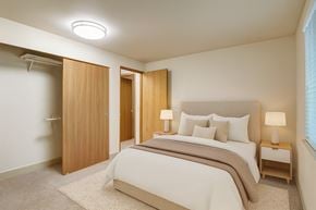 Kent apartment for rent - A calm bedroom here at Island Park featuring a bed with neutral bedding and layered pillows, a soft area rug over plush carpeting, and matching bedside tables with modern lamps. The space includes a sliding closet with wood doors, light-colored walls, and a wide window with blinds that brings in natural light.