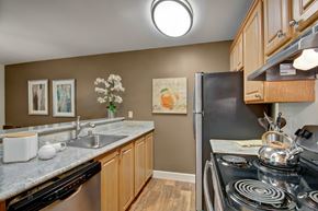 Kent apartment for rent - A functional kitchen here at Island Park featuring warm wood cabinetry, granite-style countertops, and a stainless-steel sink with a modern faucet. The space includes a full appliance suite, wood-look flooring, and coordinated wall art above the counter, creating a clean and practical layout with a welcoming feel.