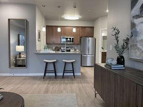 Wenatchee apartment for rent - A modern open kitchen and living area here at LIVEasy Street featuring warm wood cabinetry, a stainless-steel refrigerator and range, a breakfast bar with two upholstered stools, pendant lighting, wood-style flooring, and a neutral living space with accent décor and soft natural light creating a welcoming apartment layout.