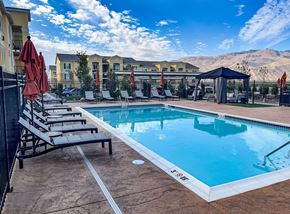 Wenatchee apartment for rent - A resort-style outdoor swimming pool here at LIVEasy Street featuring clear blue water surrounded by a stamped concrete deck, rows of cushioned lounge chairs, and closed patio umbrellas. A shaded cabana seating area sits poolside, with modern apartment buildings and scenic rolling hills visible in the background under a bright, open sky.