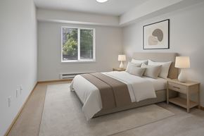 Everett apartment for rent - A calm bedroom here at Lake Park featuring a neatly made bed with light neutral bedding and accent pillows, matching bedside tables with lamps, soft carpet flooring, and a large window with blinds that brings in natural light, complemented by simple wall art and clean white walls that create a comfortable and inviting sleeping space.