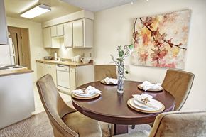 Mountlake Terrace apartment for rent - A cozy dining area here at Lakeside showcasing a round dining table set for four with upholstered chairs, neutral carpeting, and a decorative centerpiece, complemented by soft wall art and an adjacent kitchen with light cabinetry, ample counter space, and overhead lighting that creates a warm, inviting flow between spaces.