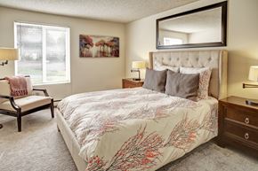 Mountlake Terrace apartment for rent - A welcoming bedroom here at Lakeside showcasing a plush bed with patterned bedding and layered pillows, flanked by matching wood nightstands with table lamps, complemented by a cozy accent chair, soft carpet flooring, wall art, and a large window with blinds that brings in natural light.