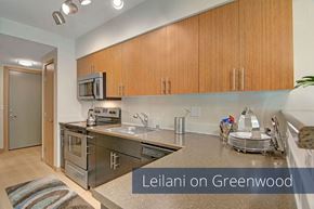 Seattle apartment for rent - A sleek kitchen here at Leilani on Greenwood featuring flat-panel wood cabinetry, smooth countertops, and a stainless steel sink set beneath under-cabinet lighting. Dark appliances line the workspace, while light wood-style flooring and modern fixtures create a clean, contemporary cooking area.