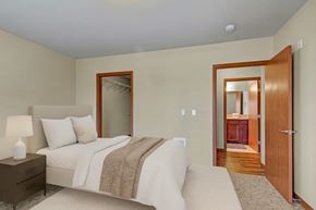 Kirkland apartment for rent - A calm bedroom here at Ondine at Juanita Bay featuring a light upholstered bed with white bedding and a neutral throw, a wood nightstand with table lamp, soft carpet flooring, and light walls, with an open closet visible and a doorway leading to a bathroom with wood cabinetry, creating a simple and comfortable retreat.