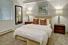 Everett apartment for rent - A serene bedroom here at Portsmith featuring a neatly made bed with crisp white bedding and patterned accent pillows, flanked by matching nightstands with tall table lamps, complemented by soft carpet flooring, a large wall mirror, abstract artwork above the headboard, and a window with blinds that brings in natural light.
