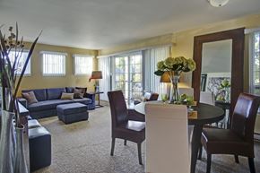 Renton apartment for rent - A spacious living and dining area here at Regency Woods featuring plush carpet flooring, a sectional sofa with an ottoman, a round dining table with mixed upholstered chairs, large windows with vertical blinds, and a sliding glass door leading to a private balcony, creating a warm, open layout filled with natural light.