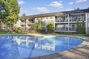 Renton apartment for rent - A peaceful outdoor swimming pool here at Regency Woods showcasing a curved pool with calm blue water, a surrounding concrete deck, and a black metal fence, set in front of multi-story apartment buildings with private balconies, mature trees, landscaped greenery, and a bright blue sky creating a relaxed community atmosphere.