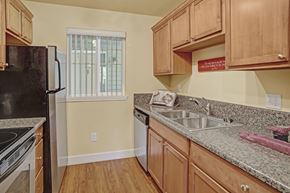 Renton apartment for rent - A cozy kitchen here at Regency Woods showcasing warm wood cabinetry, speckled countertops, and a double-basin sink beneath upper cabinets, with a dishwasher, refrigerator, and range arranged along the walls, wood-style flooring underfoot, and a window with vertical blinds bringing in soft natural light.