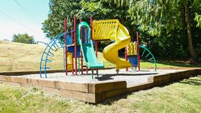 Renton apartment for rent - A colorful outdoor playground here at Regency Woods featuring a raised play structure with red support posts, a green straight slide, a yellow spiral slide, climbing ladders, and activity panels, all set on a gravel play surface bordered by timber edging and surrounded by open grassy areas and mature evergreen trees.