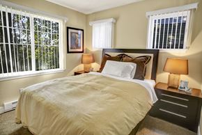Renton apartment for rent - A cozy bedroom here at Regency Woods featuring a comfortable bed with layered neutral bedding, matching nightstands with warm table lamps, soft carpeted flooring, and multiple windows with vertical blinds that allow natural light while offering leafy outdoor views.