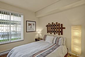 Wenatchee apartment for rent - A cozy bedroom here at Riverside 9 with a comfortable bed, warm wood headboard accent, bedside table with lamp, and a large window with vertical blinds bringing in natural light.