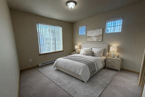 Moses Lake apartment for rent - A spacious bedroom here at The Vintage with soft beige carpet, light neutral walls, and a large window with vertical blinds bringing in natural light. A bed with white bedding and neutral accent pillows sits between matching nightstands with warm lamps.