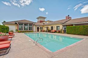 Moses Lake apartment for rent - A refreshing outdoor pool here at The Vintage with clear blue water, surrounding lounge chairs with red cushions, and a spacious patio area. A clubhouse with large windows and landscaped greenery creates a relaxing resort-style setting.