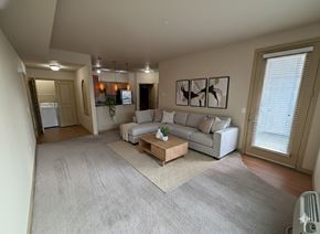 Moses Lake apartment for rent - A spacious living room here at The Vintage with soft beige carpet, warm neutral walls, and a light gray sectional sofa with accent pillows. A wooden coffee table sits on a patterned rug, while a glass door with blinds leads outside.