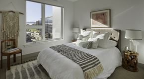 Kirkland apartment for rent - A bedroom here at Uptown at Kirkland Urban featuring bright white bedding, a soft taupe throw, warm wood nightstands, and a cozy textured rug, creating a calm, inviting space with beautiful natural light and window views.