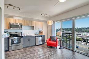 Seattle apartment for rent - A stylish open kitchen and living area here at Vive featuring modern cabinetry, stainless steel appliances, a vibrant red accent chair, warm wood-tone flooring, and large sliding glass doors opening to a bright private balcony with city views.