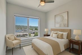 Seattle apartment for rent - A bright bedroom here at Vive featuring soft neutral bedding, warm wood nightstands, a cozy accent chair, and a large window that fills the inviting space with natural light and scenic neighborhood views.