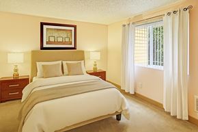 Lynnwood apartment for rent - A bright bedroom here at Whispering Cedars with beige carpet, cream walls, and large windows with white curtains, featuring a comfortable bed with neutral bedding, matching bedside tables with lamps, and a warm, inviting atmosphere.
