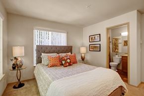 Issaquah apartment for rent - A cozy bedroom here at Windsong featuring a tufted headboard bed, orange accent pillows, and matching bedside lamps. Bright, welcoming layout with soft carpet, neutral tones, and convenient ensuite bathroom access.