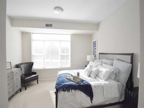 Shorewood apartment for rent - A bedroom here at Oaks of Shorewood featuring a bright layout, plush white bedding, a dark accent chair, large sunlit window, and soft carpeting, creating a calm, cozy, and inviting retreat.