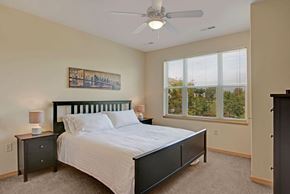 Sun Prairie apartment for rent - A bedroom here at Van Buren Place featuring a dark wood bed frame, soft carpet flooring, large bright windows with peaceful views, and a clean, cozy layout that feels warm and inviting.