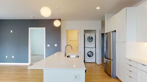 Milwaukee apartment for rent - A modern open kitchen here at Vim and Vigor with a large island and sink, white cabinets, stainless steel refrigerator, pendant lighting, wood-style flooring, and an in-unit stacked washer and dryer nearby.