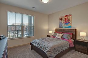 Fitchburg apartment for rent - A bright bedroom here at Vue at Pinnacle Park with soft carpet, a large window with blinds and neighborhood views, a wood bed frame with gray and pink bedding, and matching nightstands with warm bedside lighting.
