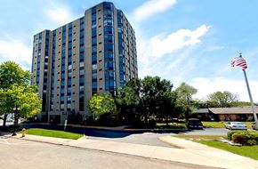 Pekin apartment for rent - A tall building with a flag on top is surrounded by trees and a street in front.