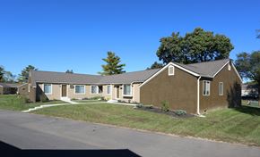 Columbus apartment for rent - A brown house with a white door and windows is surrounded by a grassy area.