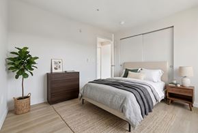 Seattle apartment for rent - Staged bedroom with a bed, nightstand, dresser, potted plant, and sliding closet doors over light flooring.