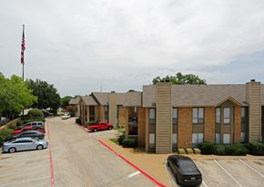Grapevine apartment for rent - A parking lot with a red car and a black car in front of a building with a flag on top.
