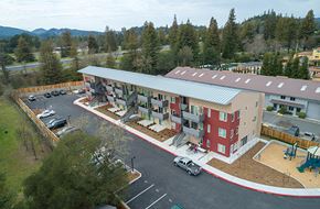 Healdsburg apartment for rent - A large building with a red and white exterior is surrounded by a parking lot and trees.