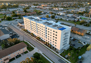Cape Coral apartment for rent - A large white building with a blue roof is surrounded by a parking lot and other buildings.